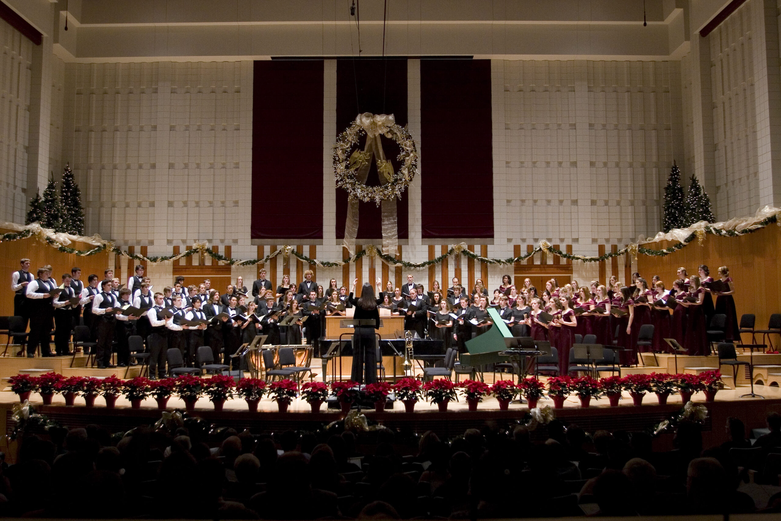 Three choirs, voce Femminile, Male Ensemble Willamette and Chamber choir, sing together in Hudson Hall, which is decorated with a large wreath, lit Christmas trees, and a row of poinsettias.